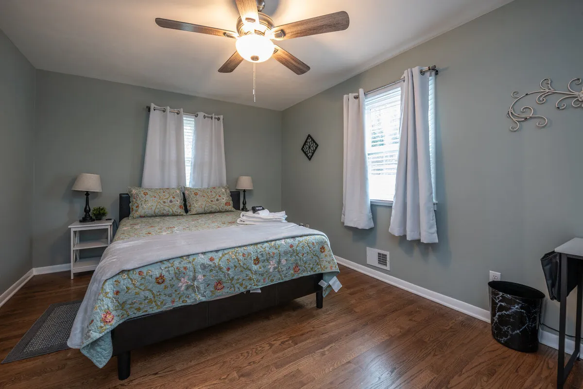 A calm and cozy bedroom with a comfortable bed, nightstand, lamp, and large windows bathing the space in natural light. The soothing palette and simple, thoughtful decor emphasize a homey retreat ideal for a restful night or quiet moments spent reading by the window.