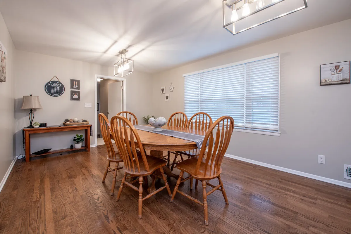 The dining area features a wooden dining table surrounded by matching wooden chairs, with a lamp above and decorative wall items that create a cozy, welcoming atmosphere. The table is set for gatherings, suggesting both casual meals and festive occasions.
