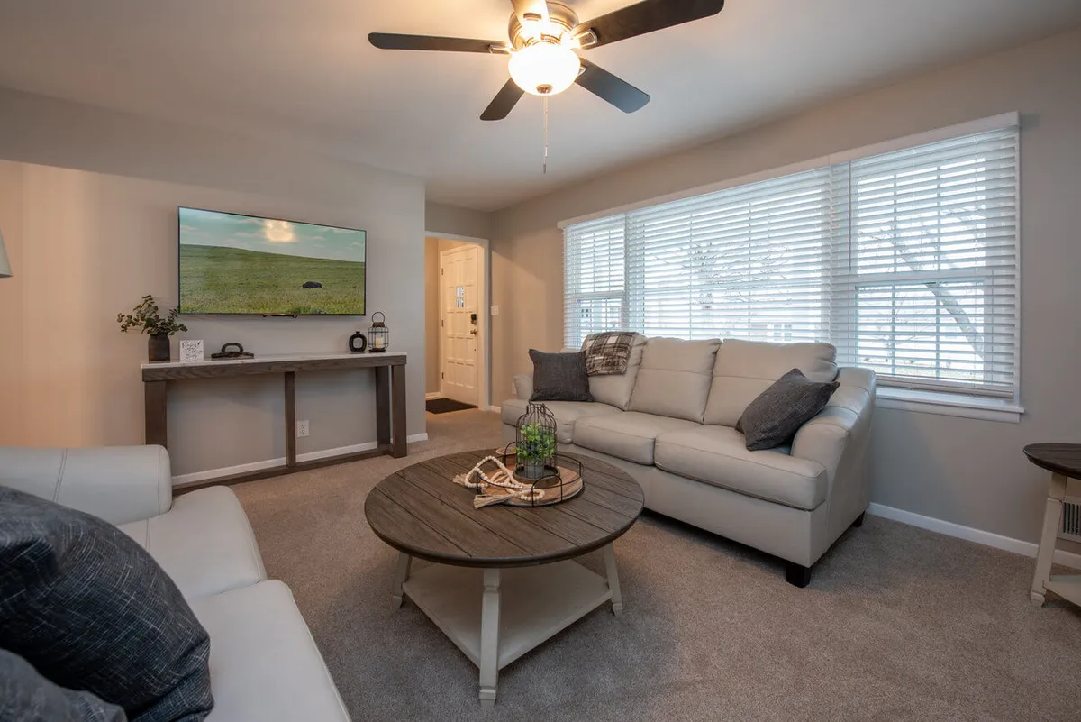 A welcoming living space showcasing a sofa and coffee table set beneath a large window, with a TV and console table for entertainment. The layout is open and designed to foster social interaction and relaxation.