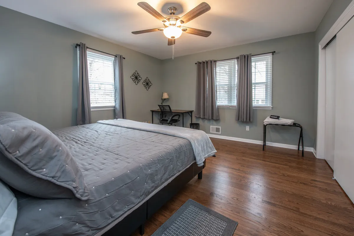 A cozy bedroom setup featuring a bed near large windows, a desk for working or planning, and a serene atmosphere that suggests a peaceful retreat after a day out. Natural light pours in, highlighting the tranquil ambiance and the possibility of relaxing with a morning coffee or catching up on emails.