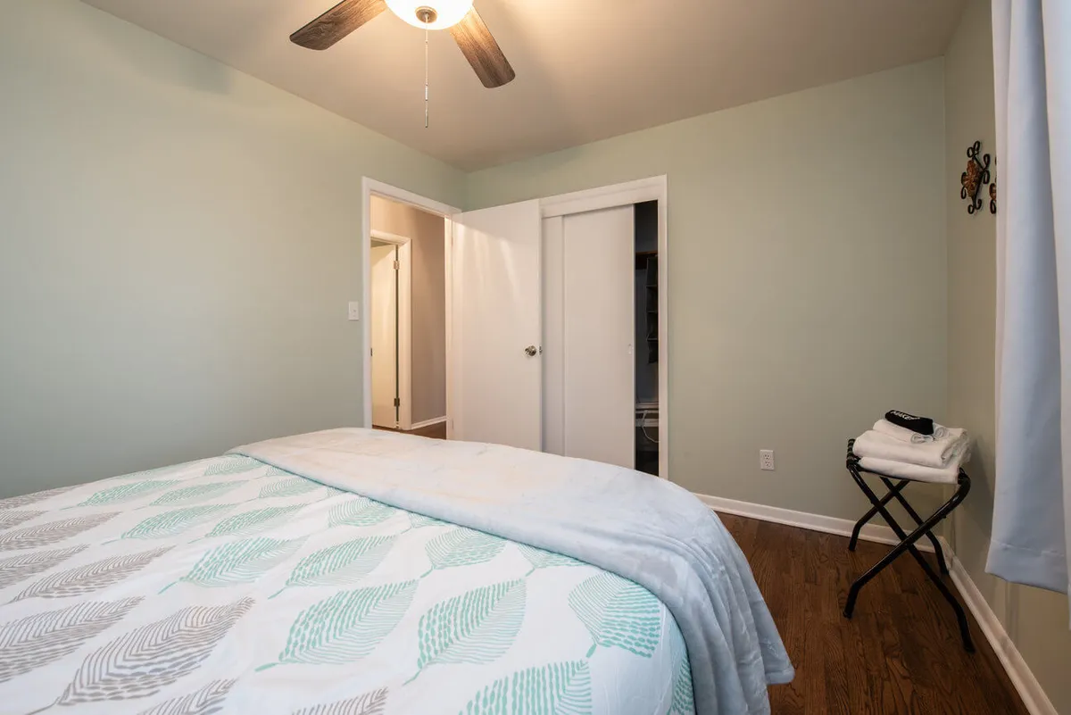 A serene bedroom featuring a bed with patterned bedding, a ceiling fan overhead, and natural light streaming through the windows. Doorways offer a sense of privacy and comfort, while soft furnishings and gentle lighting create a tranquil atmosphere ideal for relaxation or unwinding with a book.